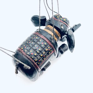 May include: A black wooden puppet of an elephant wearing a red, yellow, and black patterned armor. The puppet is suspended by strings and has a long trunk.