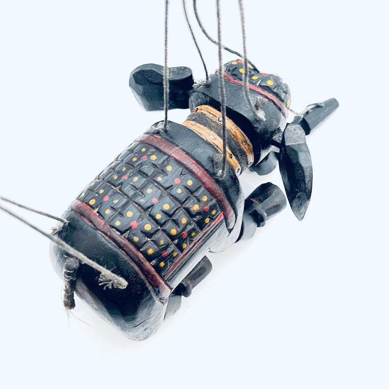 May include: A black wooden puppet of an elephant wearing a red, yellow, and black patterned armor. The puppet is suspended by strings and has a long trunk.