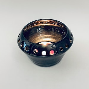 Puede incluir: Un portavelas de cerámica negro con un borde dorado y agujeros alrededor del borde. Una pequeña vela blanca está ardiendo dentro del portavelas.