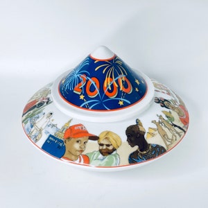 Könnte beinhalten: Eine weiße Keramik-Schale mit Deckel, verziert mit einem blauen und roten Design, das das Jahr 2000 und Feuerwerk zeigt. Die Schale hat eine farbenfrohe Szene mit Menschen und Tieren am Rand.