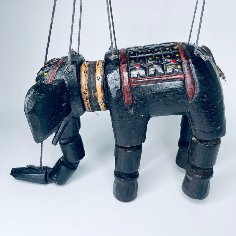 May include: A black wooden puppet of an elephant with red and yellow painted details. The puppet is suspended by strings and has a movable head, legs, and trunk.