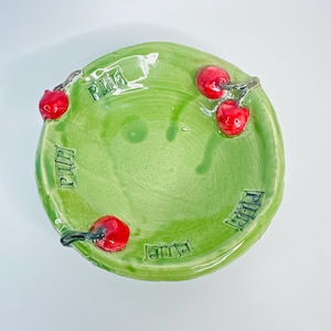 May include: A handmade ceramic bowl in a vibrant green color, adorned with four red cherry accents. The bowl features the word "Rima" in a stylized font around the rim. The cherries have a glossy finish, adding a touch of realism to the design.