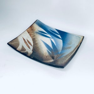 Può includere: Un piatto da portata quadrato in ceramica con una glassa blu, marrone e bianca. Il piatto ha una curva sottile e presenta un design stilizzato di foglie.