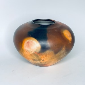 Puede incluir: Un jarrón de cerámica hecho a mano con cuerpo redondeado y abertura oscura. El jarrón presenta una mezcla de tonos marrón, negro y naranja, con patrones circulares más claros. El diseño es orgánico y terroso.