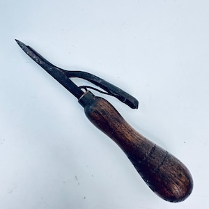 May include: An antique metal tool with a wooden handle. The tool has a pointed end and a curved metal piece. The handle is dark brown and appears to be made of wood. The metal parts are dark and show signs of age.