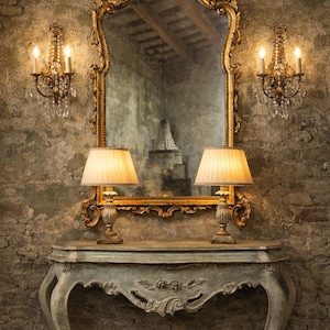 Peut inclure: Un miroir doré orné est suspendu au-dessus d'une console grise, flanqué de deux appliques et de deux lampes. Le miroir reflète une pièce avec une structure décorative. Le mur est en briques vieillies.