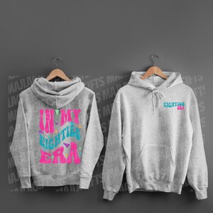 Puede incluir: Dos sudaderas con capucha grises. Una tiene el texto "In My Eighties Era" en letras rosa y azul con triángulos de colores. La otra tiene el texto "Eighties Era" en letras azules.