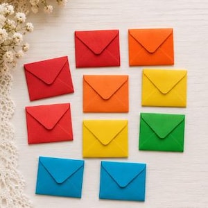 Pode incluir: Uma coleção de dez pequenos envelopes de papel quadrados dispostos em uma grade. Os envelopes apresentam um design clássico de aba triangular e vêm em um vibrante espectro de arco-íris de cores sólidas, incluindo vermelho, laranja, amarelo, verde e azul.