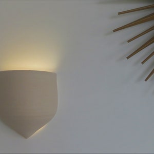 Op de afbeelding: Een beige wandlamp met een schildvorm, die een warm licht uitstraalt. Gemonteerd op een witte muur, met een gouden sterrenpatroon in de rechterbovenhoek. De lamp zorgt voor sfeervolle verlichting.