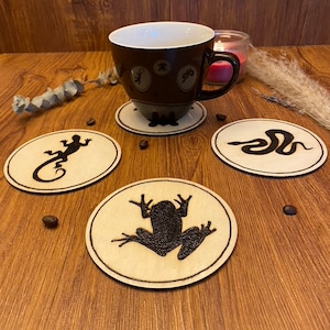 May include: A brown teacup with a white interior sits on a matching coaster. Three additional coasters with black animal silhouettes of a lizard, snake, and frog are displayed on a wooden surface. Coffee beans are scattered around.