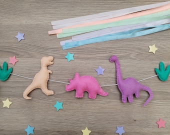 May include: A felt dinosaur banner with a Tyrannosaurus Rex, Triceratops, and Brontosaurus in peach, pink, and purple. The banner includes felt stars and green fern accents. Pastel ribbons are arranged above the banner on a wooden surface.