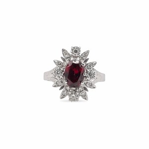 Peut inclure: Une bague en argent ornée d'une pierre précieuse rouge foncé de forme ovale au centre, entourée d'un halo de petites pierres précieuses claires. Le design de la bague rappelle une fleur ou un éclat de soleil, avec les petites pierres rayonnant vers l'extérieur.