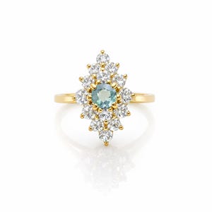 Peut inclure: Bague en or avec une pierre centrale ronde bleu clair entourée de pierres blanches. La bague présente un motif floral ou en forme d'étoile, avec un anneau en or poli.