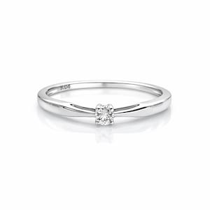 Peut inclure: Une bague en argent brillant avec un petit diamant rond au centre. L'anneau est fin et poli, avec une légère courbe. La bague est présentée sur une surface blanche.