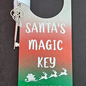 May include: Wooden door hanger with a key, featuring a white, red, and green gradient. The text reads "SANTA'S MAGIC KEY FOR OUR HOME WITHOUT A CHIMNEY" with a Santa and reindeer graphic. A silver key is attached.