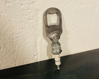 May include: A silver bottle opener made from a spark plug. The opener is attached to the spark plug with a metal ring. The spark plug is white with black lettering that says "Champion".