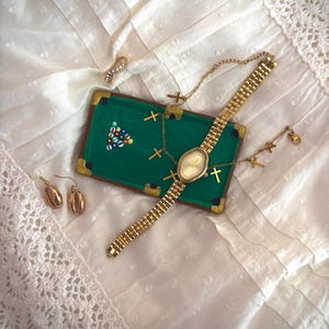 May include: A collection of gold-toned jewelry and accessories, including a wristwatch, earrings, a pendant, and a necklace with cross charms. A miniature green pool table with colorful billiard balls is also present.