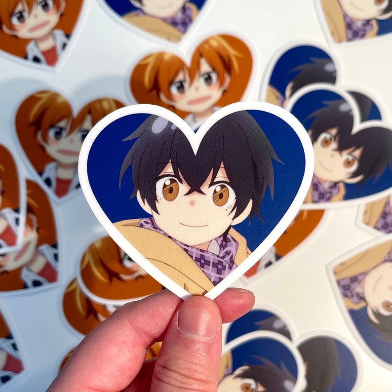 May include: A heart-shaped sticker featuring a cartoon character with brown hair, brown eyes, and a light brown jacket. The character is smiling and wearing a purple and white scarf. The background is a dark blue color.