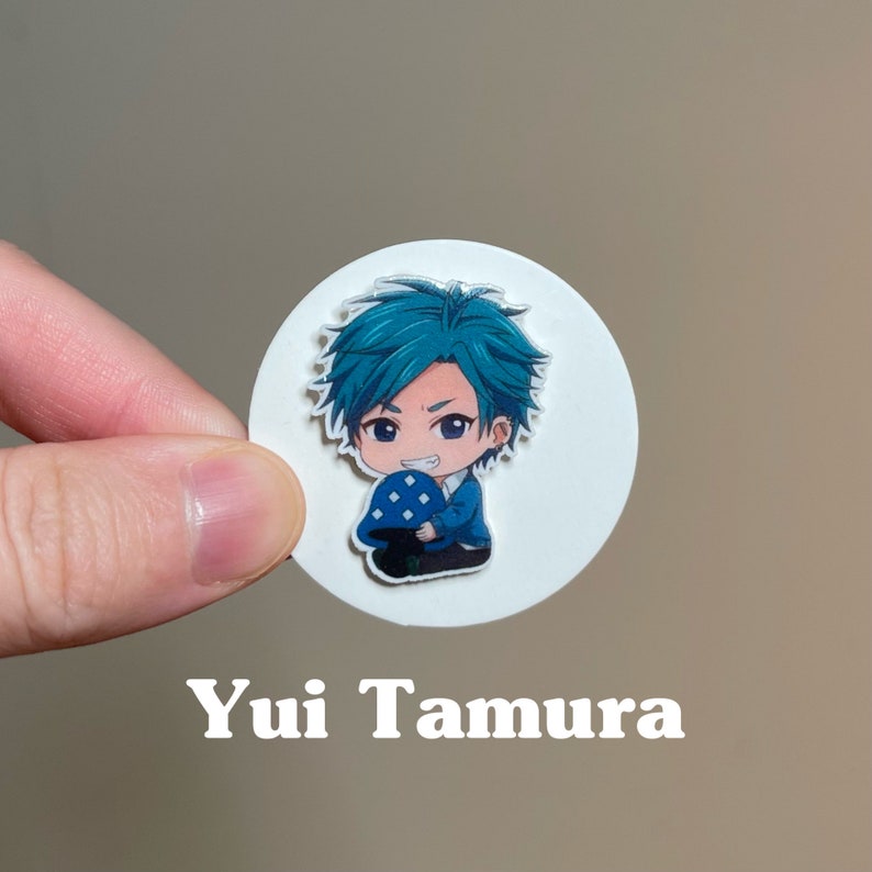 May include: A white circle sticker with a cartoon illustration of a young man with blue hair holding a blue mushroom. The text "Yui Tamura" is printed below the sticker.