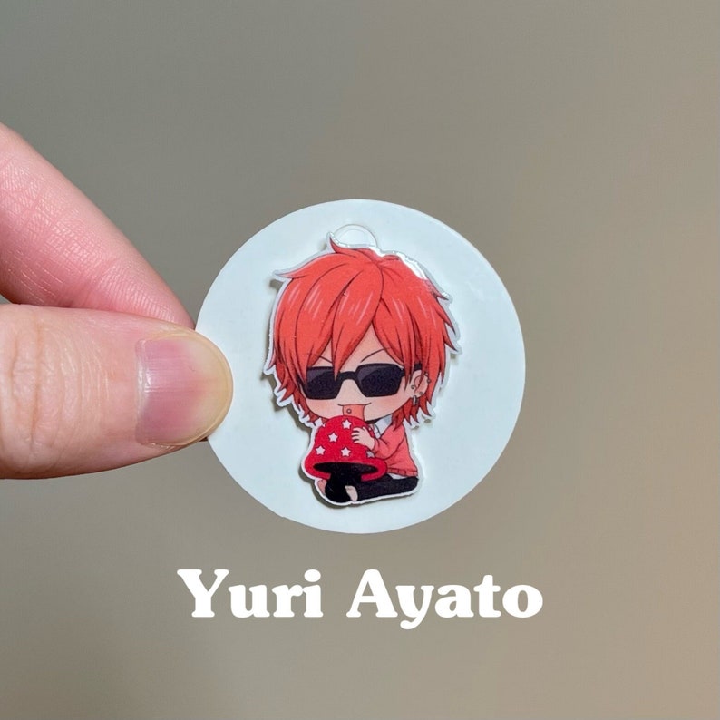 May include: A round sticker with a cartoon image of a red-haired anime character wearing sunglasses and a red sweater. The character is holding a red mushroom with white spots. The sticker has the text "Yuri Ayato" printed below the image.