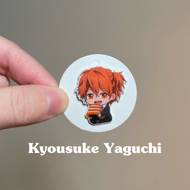 May include: A white circle with a cartoon image of a character with orange hair, wearing a black suit and holding a slice of cake. The text "Kyousuke Yaguchi" is printed below the circle.