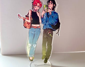 May include: Acrylic stand featuring two illustrated figures. One figure has red hair, a black top, and blue jeans, carrying a bag. The other figure, with black hair, wears a blue jacket and holds a phone. The stand is clear with a circular base.