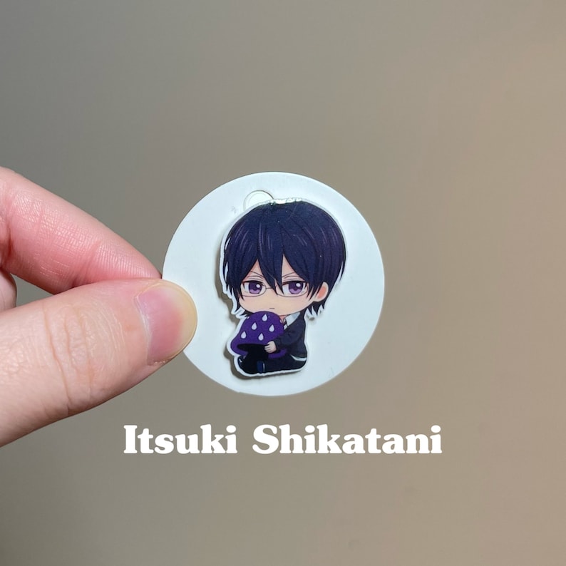 May include: A white circle with a cartoon character holding a purple mushroom. The character has black hair, glasses, and is wearing a black suit. The text "Itsuki Shikatani" is printed below the circle.
