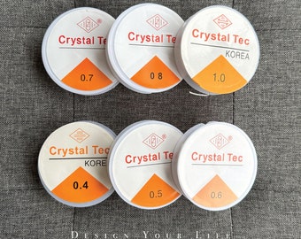 Pode incluir: Seis rolos redondos brancos de fio elástico transparente com texto vermelho e preto. Os rolos são etiquetados como "Crystal Tec" e têm tamanhos diferentes: 0,4, 0,5, 0,6, 0,7, 0,8 e 1,0.