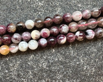 Pode incluir: Um fio de contas redondas de pedras preciosas multicoloridas. As contas são uma mistura de rosa, branco e cinza com algumas manchas pretas. As contas são enfiadas em um fio fino rosa.