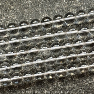 Könnte beinhalten: Eine Reihe von durchsichtigen Glasperlen, die in Reihen auf einer grauen, strukturierten Oberfläche angeordnet sind. Die Perlen sind rund und glatt, mit einem leichten Glitzern.