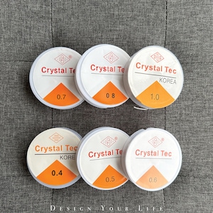 Könnte beinhalten: Sechs weiße Spulen mit transparentem elastischem Faden, beschriftet mit "Crystal Tec" in verschiedenen Größen: 0,4, 0,5, 0,6, 0,7, 0,8 und 1,0.