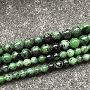 Könnte beinhalten: Eine Reihe grüner Edelsteinperlen mit schwarzen und weißen Einschlüssen. Die Perlen sind rund und glatt und in einer Reihe auf einem grauen Hintergrund angeordnet.