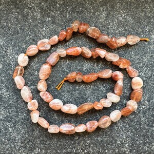 Pode incluir: Um fio de contas de pedra do sol em forma de seixos. As contas são uma mistura de pêssego claro e laranja com uma qualidade brilhante e iridescente.
