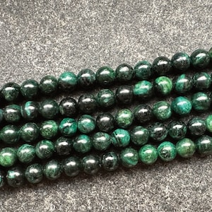 Op de afbeelding: Een streng donkergroene edelsteen kralen. De kralen zijn rond en hebben een gevlekt, gemarmerd uiterlijk.