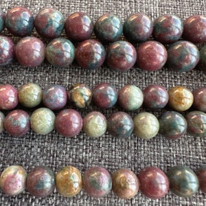 Pode incluir: Um fio de contas redondas de pedras preciosas multicoloridas. As contas são uma mistura de verde, vermelho e marrom com um padrão salpicado.