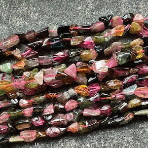 May include: A strand of multi-colored, faceted, and tumbled gemstone beads. The beads are a mix of pink, green, brown, and black. The beads are strung on a pink thread.