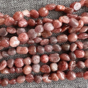 Op de afbeelding: Een streng roze en rode edelsteen kralen, elke kraal is een gladde, afgeronde ovale vorm. De kralen zijn in een losse stapel op een getextureerd grijs oppervlak gerangschikt.