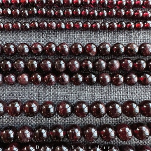 Op de afbeelding: Meerdere strengen donkerrode edelsteen kralen, waarschijnlijk granaten, gerangschikt in rijen op een grijze stoffen achtergrond. De kralen zijn glad en rond, met een lichte variatie in grootte.