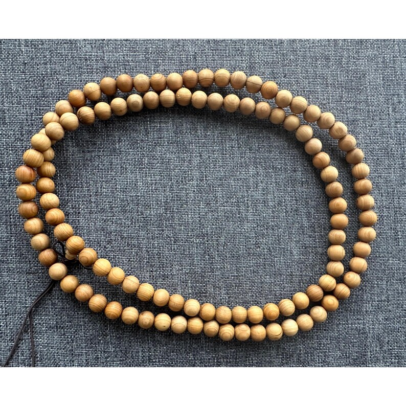 Puede incluir: Un collar de cuentas de madera de doble hebra. Las cuentas son de color marr&oacute;n claro y esf&eacute;ricas, ensartadas en un cord&oacute;n oscuro. El collar est&aacute; colocado sobre una superficie gris texturizada.