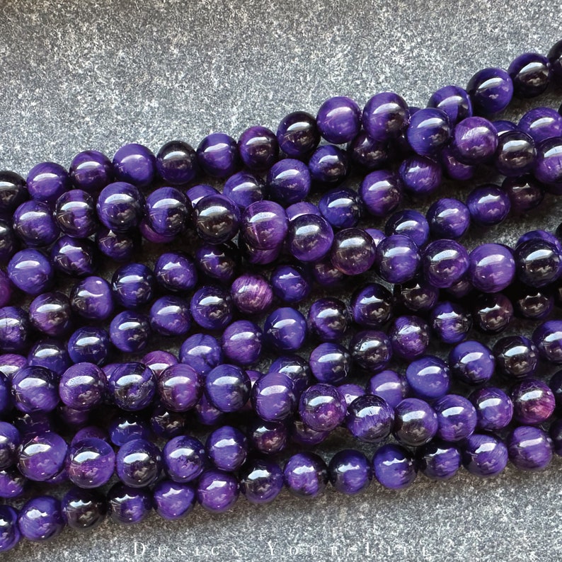 May include: Strands of polished, round purple beads. The beads have a glossy finish and reflect light, creating highlights. The beads are a deep, rich purple color, with some variation in tone. The image is of a collection of beads, likely for jewelry making.