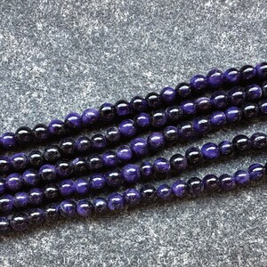 May include: Close-up of strands of round beads in shades of deep purple and black. The beads are smooth and shiny, with a polished appearance. The beads are strung together, creating a necklace or bracelet.