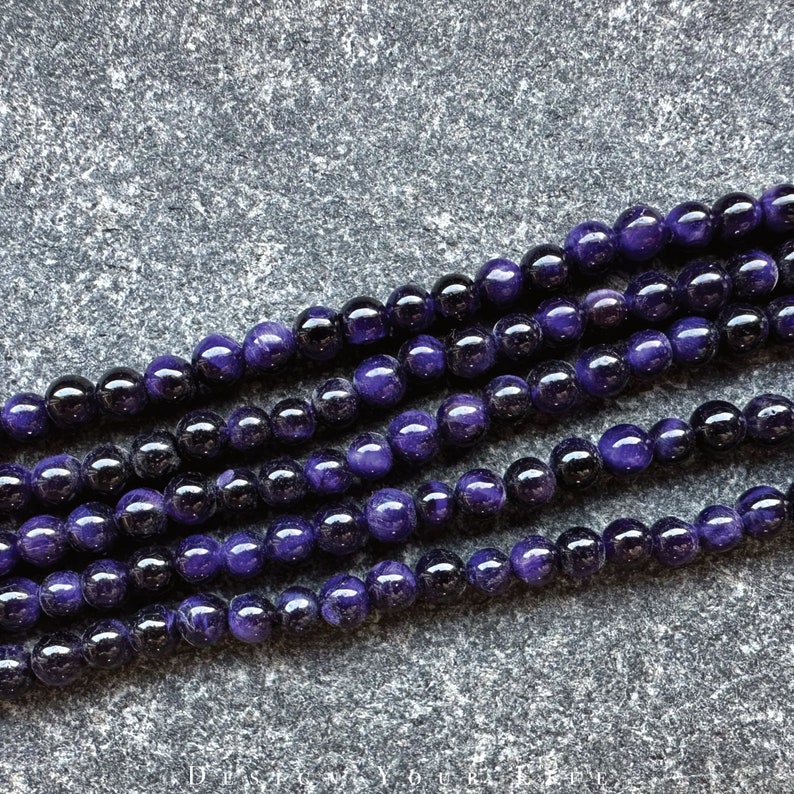 May include: Close-up of strands of round beads in shades of deep purple and black. The beads are smooth and shiny, with a polished appearance. The beads are strung together, creating a necklace or bracelet.