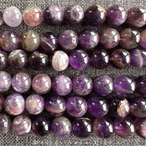 May include: A strand of round, purple amethyst beads with a smooth, polished finish. The beads have a natural, cloudy appearance with streaks of white and purple.