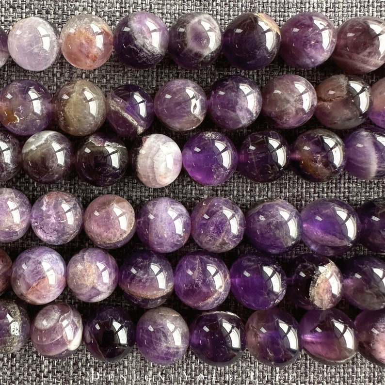 May include: A strand of round, purple amethyst beads with a smooth, polished finish. The beads have a natural, cloudy appearance with streaks of white and purple.