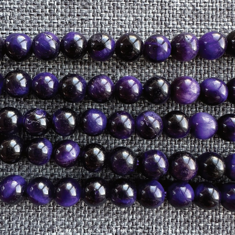 May include: Close-up of strands of polished gemstone beads. The beads alternate between deep purple and black, creating a striking contrast. The round beads are arranged in neat rows, showcasing their glossy surfaces and natural variations.