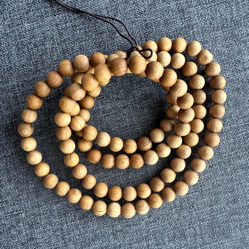 Puede incluir: Un collar de cuentas de madera marr&oacute;n claro, probablemente s&aacute;ndalo, dispuestas en m&uacute;ltiples bucles. Las cuentas esf&eacute;ricas muestran un veteado natural de la madera. Una cuerda marr&oacute;n oscuro es visible en la parte superior, uniendo las cuentas. Las cuentas son de color natural.