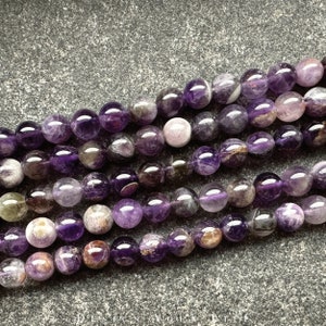 May include: A strand of amethyst beads with a mix of purple and white tones. The beads are round and have a smooth, polished finish.