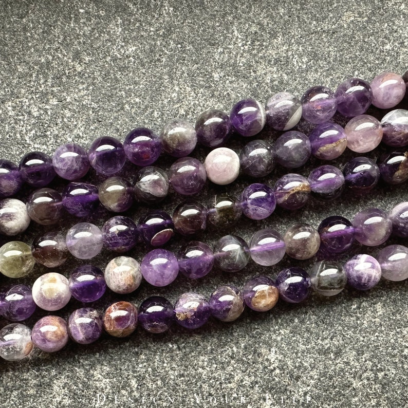 May include: A strand of amethyst beads with a mix of purple and white tones. The beads are round and have a smooth, polished finish.