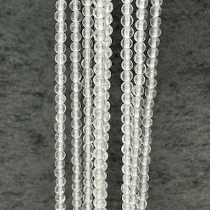 May include: Several strands of clear, round beads, possibly quartz or crystal, are shown against a grey backdrop. The beads are consistent in size and strung together, producing a delicate, shimmering effect. Suitable for jewellery making or craft projects.