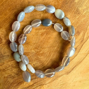 May include: A necklace of oval, polished agate beads in grey, white, and pale green. The beads are strung together, creating a smooth design. The necklace is displayed on a wooden surface. Each bead is approximately 1.5 cm in length.